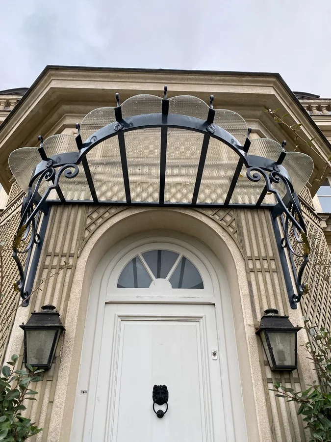 Marquise ouvragée en fer forgé au-dessus d'une porte cintrée encadrée de lanternes, L'Art de Faire