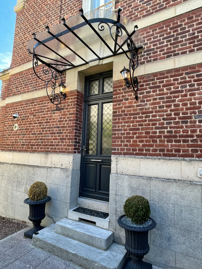 Marquise en fer forgé au-dessus d'une porte, façade en brique et topiaires, L'Art de Faire