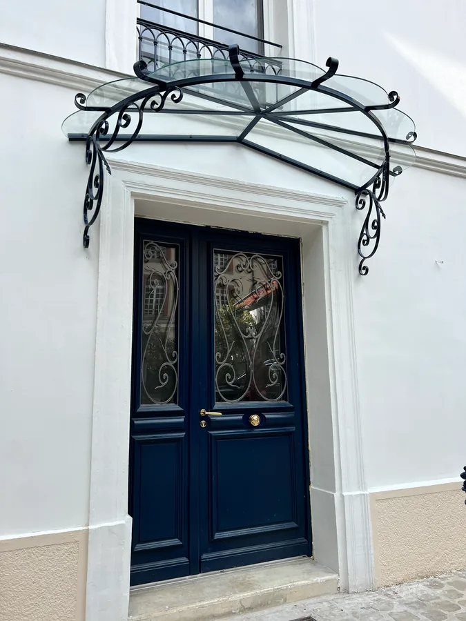 Marquise ouvragée en fer forgé à volutes au-dessus d'une porte bleue, L'Art de Faire