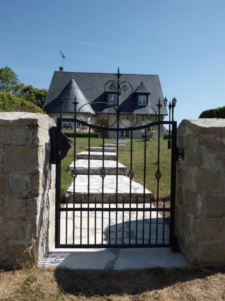 Portillon en fer forgé à barreaux et volutes entre piliers en pierre, L'Art de Faire
