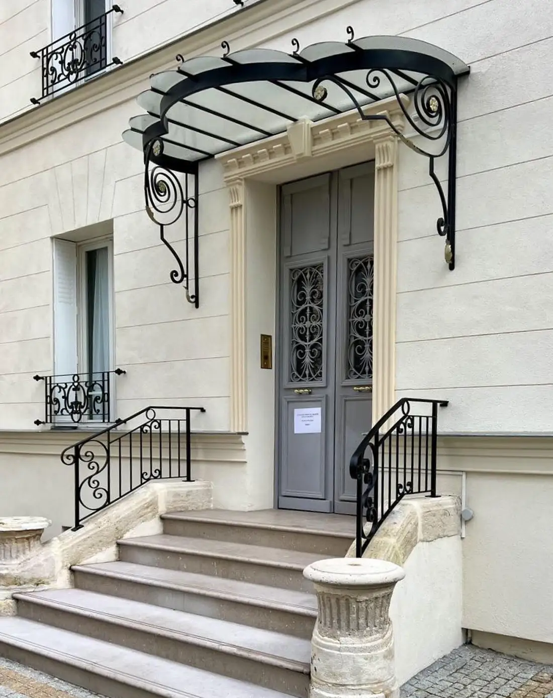 Marquise noire ornée au-dessus d'une porte d'entrée grise avec des escaliers en pierre et des balustrades en fer forgé